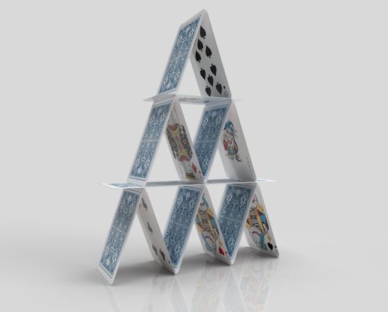 A structure composed of playing cards arranged in a triangular formation, resembling a house of cards. The cards feature various designs and suits, including spades and face cards, standing on a reflective surface against a plain background.